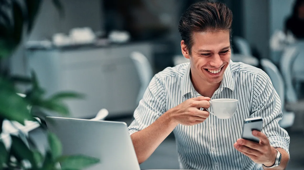 Uśmiechnięty mężczyzna z telefonem i kawą przy laptopie